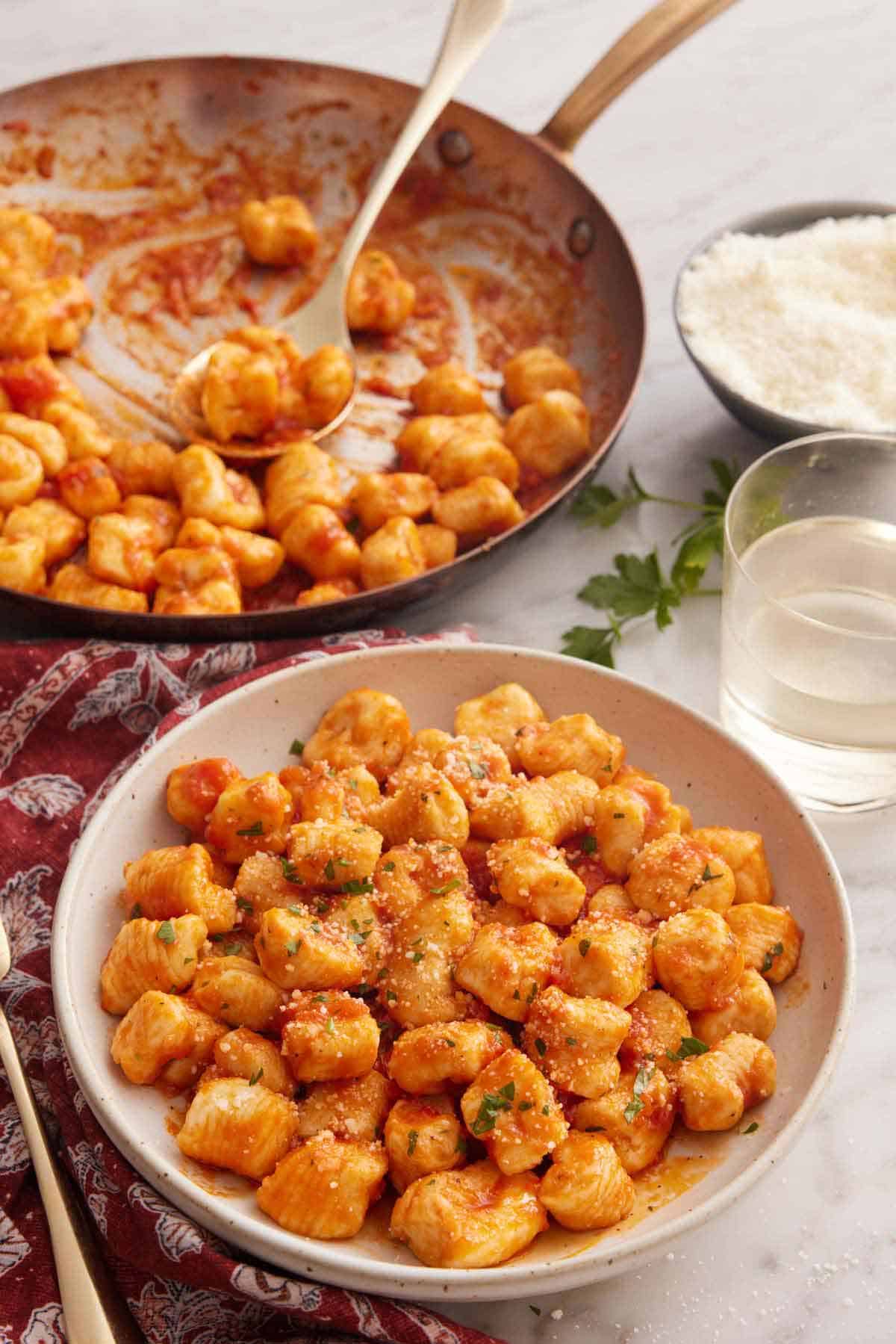 A skillet and plate of gnocchi coated in a red sauce. Glass of wine and bowl of shredded cheese in the background.