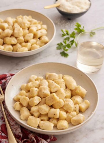 Two plates of gnocchi with a glass of wine and bowl of cheese.