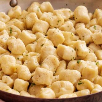 A close up view of a skillet of cooked gnocchi garnished with black pepper and chopped herbs..