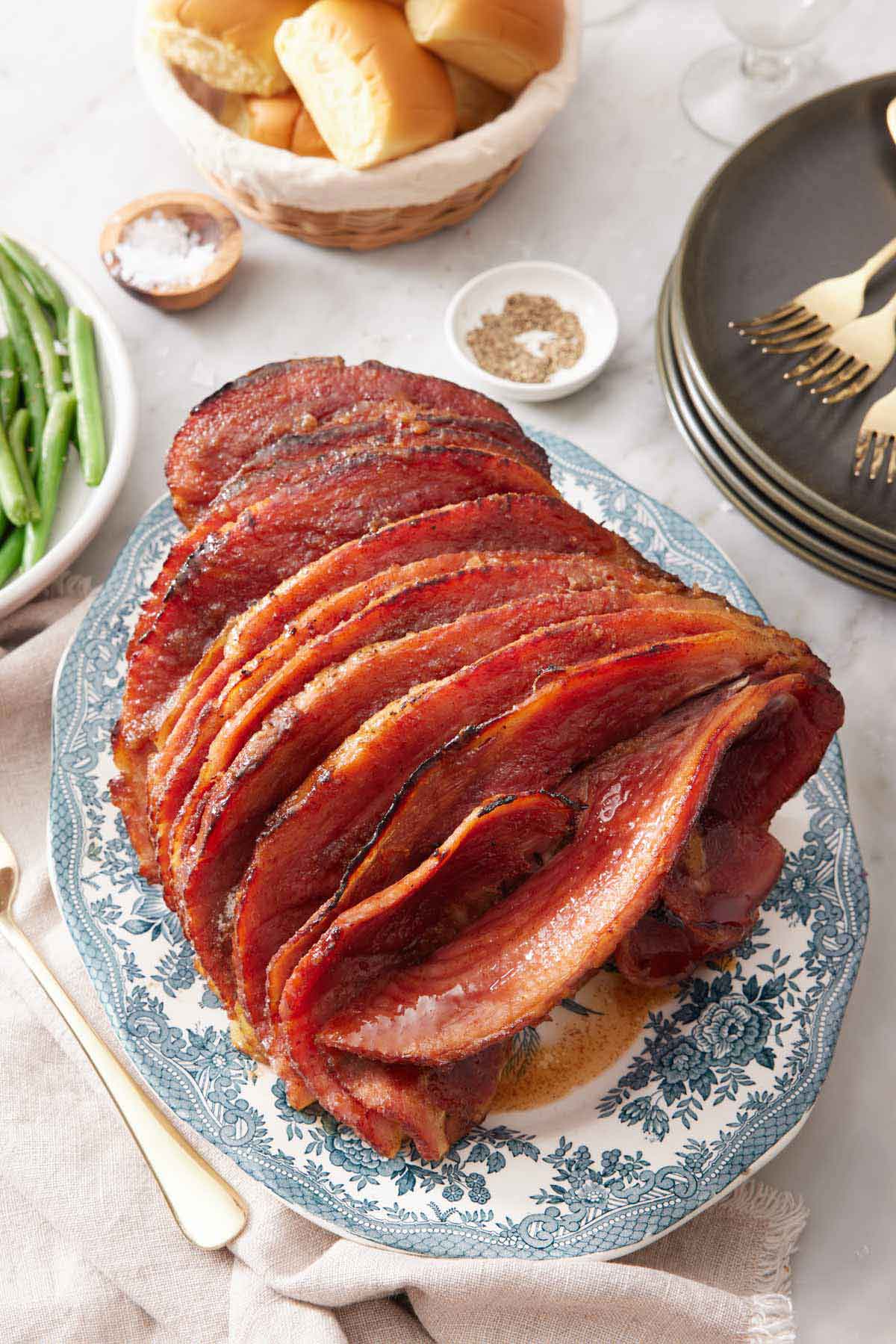 A platter with a honey baked ham with a stack of plates, bowl of bread, and some green beans off to the side.