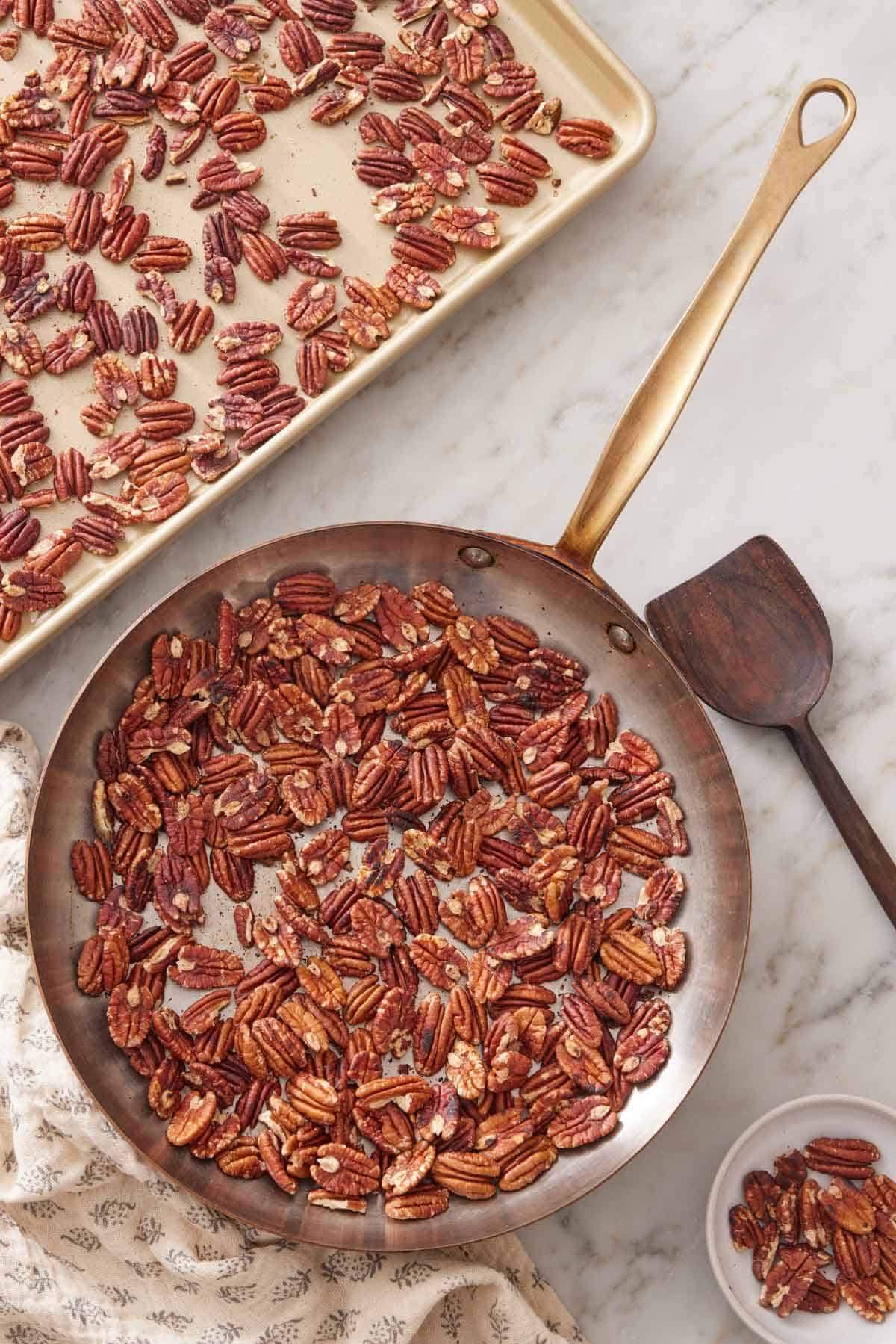 Overhead view of a skillet of toasted pecans. A wooden spoon and sheet pan of pecans off to the side.
