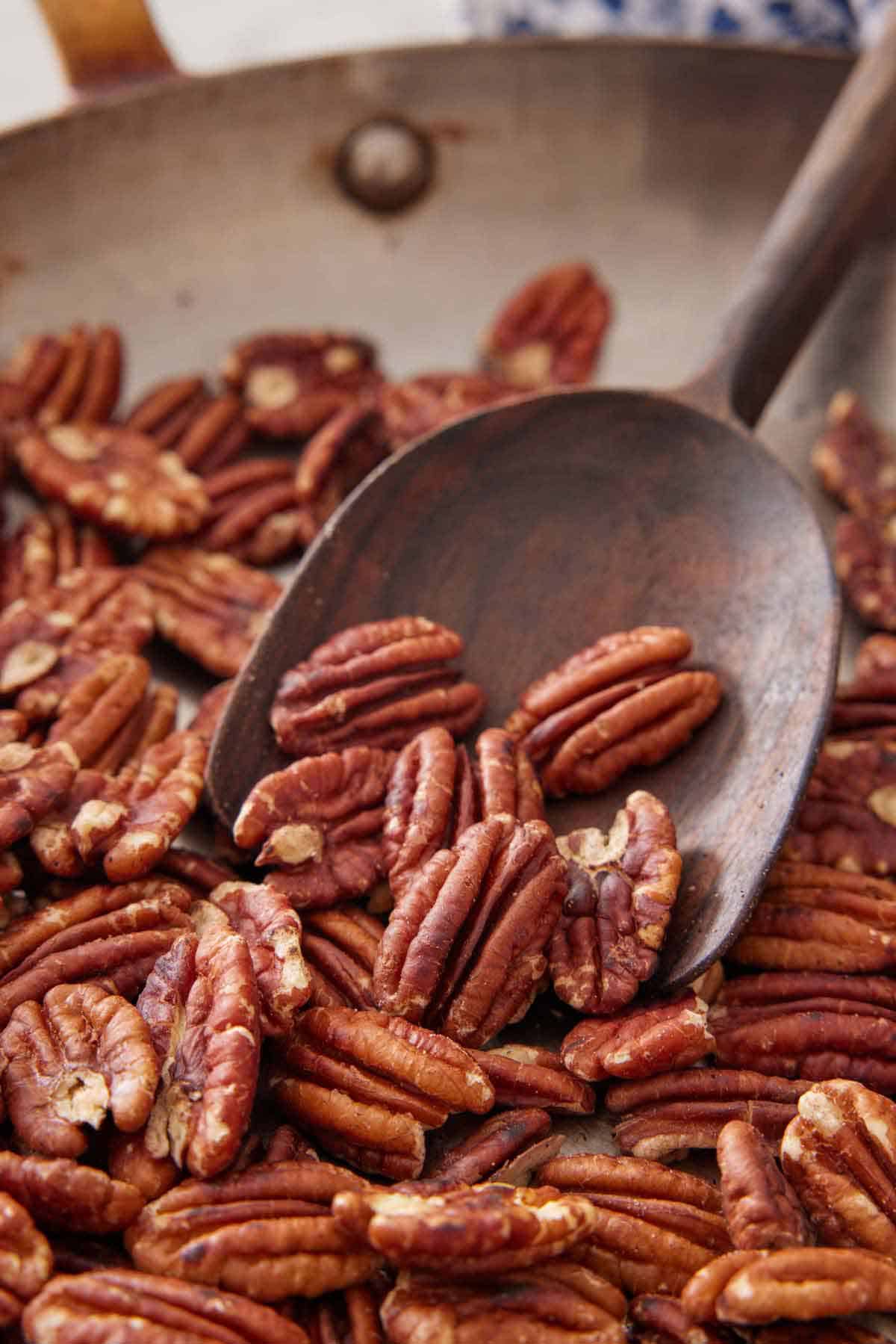 A wooden spoon scooping up some toasted pecans from a skillet.