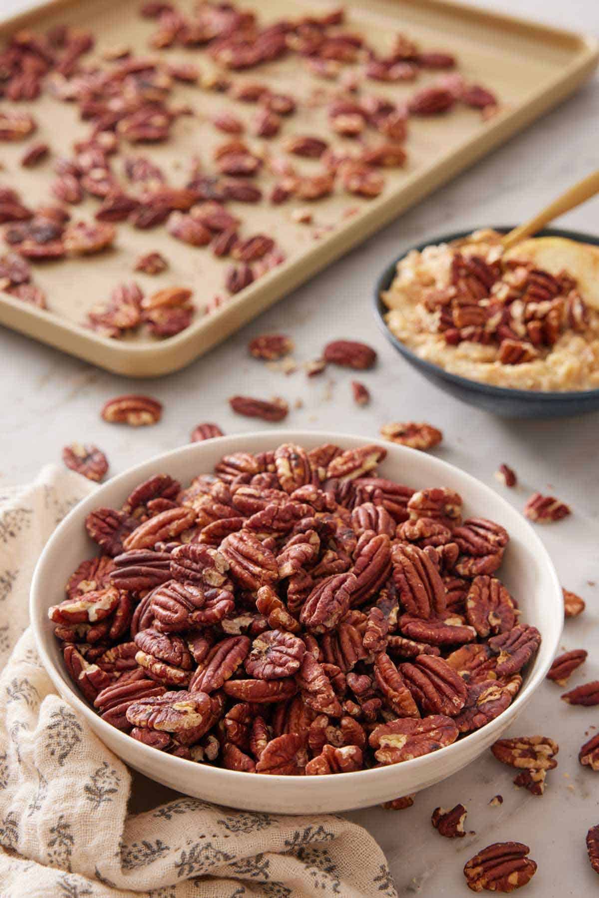 A bowl of toasted pecans with a sheet pan in the background and more pecans in a bowl over oatmeal.