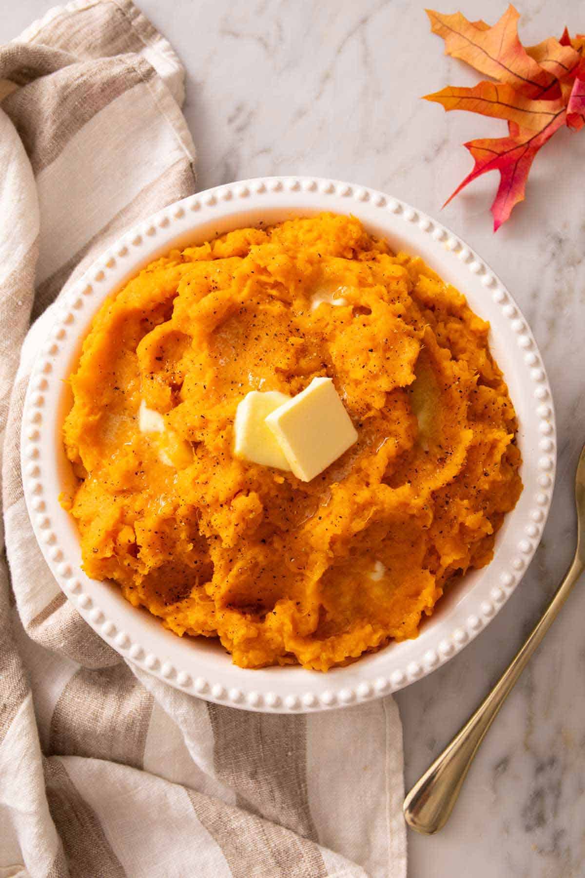 Overhead view of a bowl of mashed sweet potatoes with knobs of butter on top. Linen napkin and some leaves on the side.