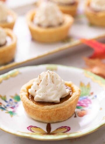 A plate with a mini pumpkin pie topped with whipped cream. More pies out of focus in the background.
