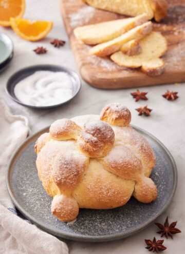 A loaf of Pan de Muerto with a wooden serving board in the back with sliced bread.