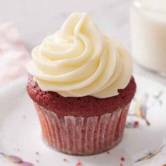 A plate with a red velvet cupcake topped with a generous helping of cream cheese frosting. Glass of milk in the back.