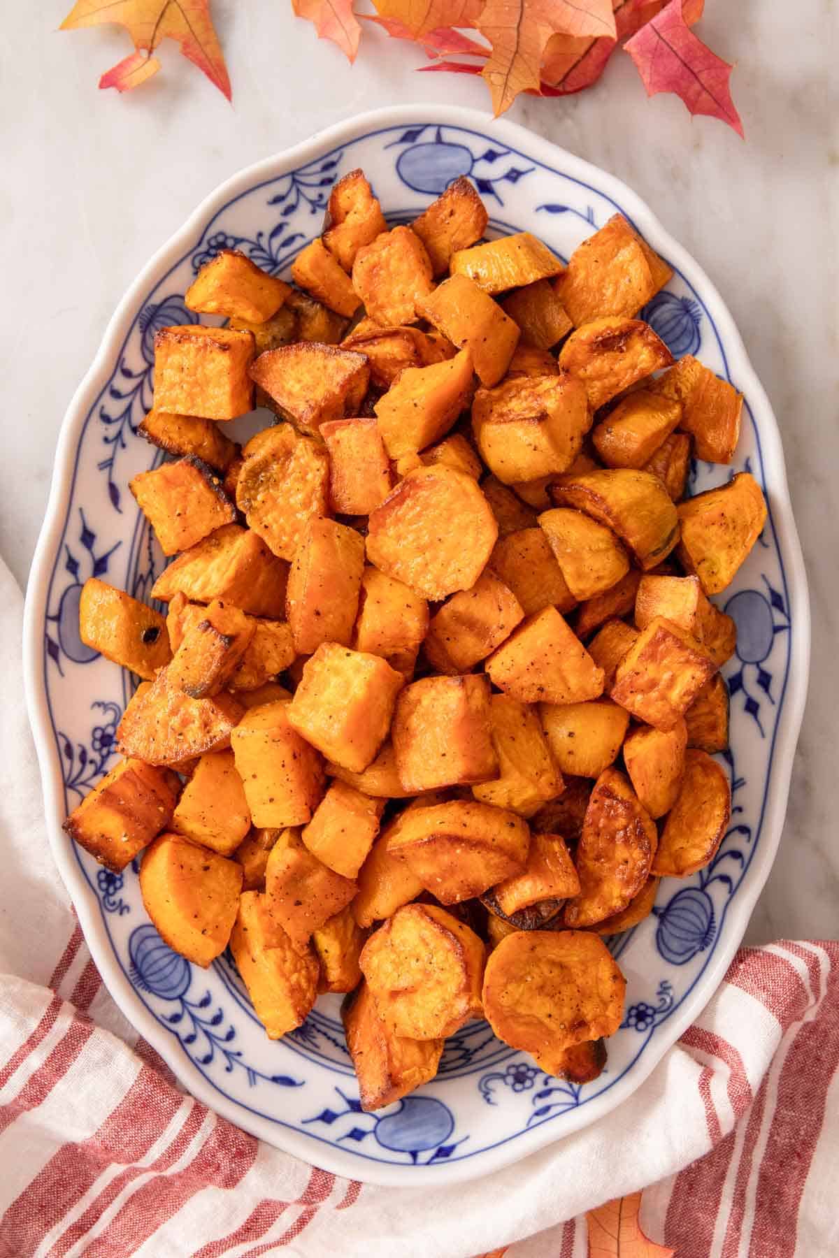 Overhead view of an oval platter of roasted sweet potatoes. Leaves and a linen surrounding it.