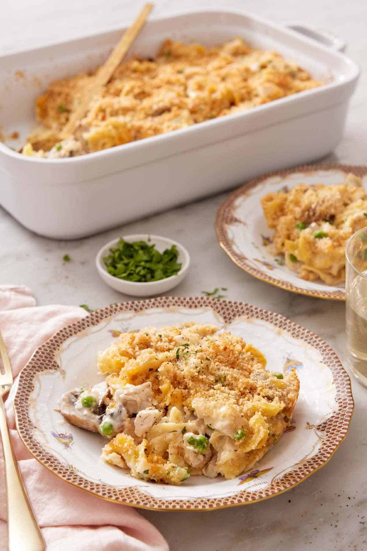 A plate of turkey casserole with a baking dish, bowl of parsley, and a second plated serving in the background.