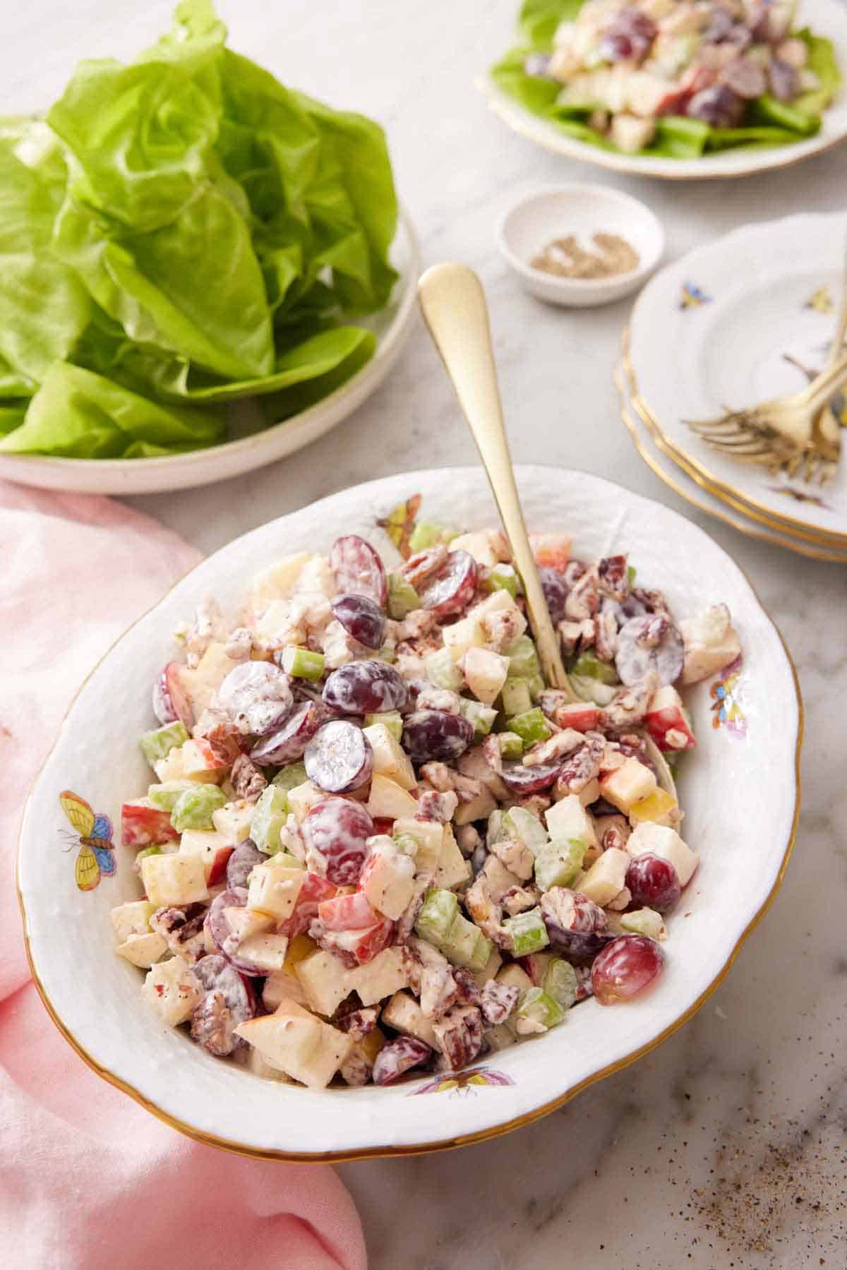 A platter of Waldorf salad with a serving spoon tucked in with some lettuce and stacked plates in the background.