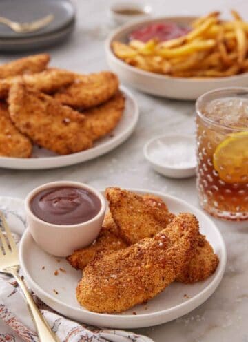 A plate with air fryer chicken tenders on a plate with bbq sauce. More tenders, some french fries, and a glass of lemon iced tea in the background.