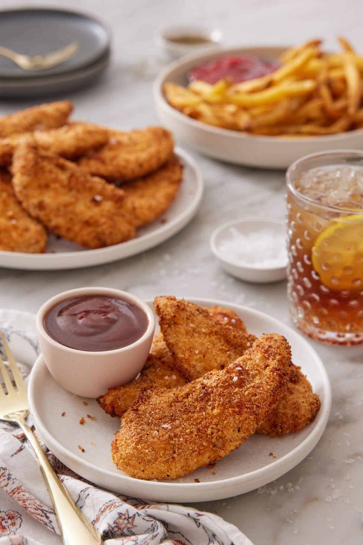 A plate with air fryer chicken tenders on a plate with bbq sauce. More tenders, some french fries, and a glass of lemon iced tea in the background.
