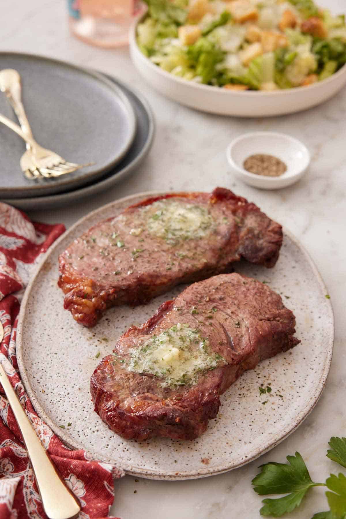 A plate with two pieces of air fryer steak topped with melted herb butter. A salad in the background along with a stack of plates and forks.