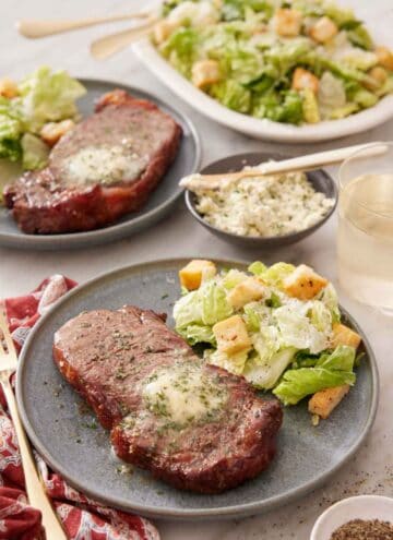 A plate with an air fryer steak with a side of salad. An additional plated serving in the background along with a platter of salad.