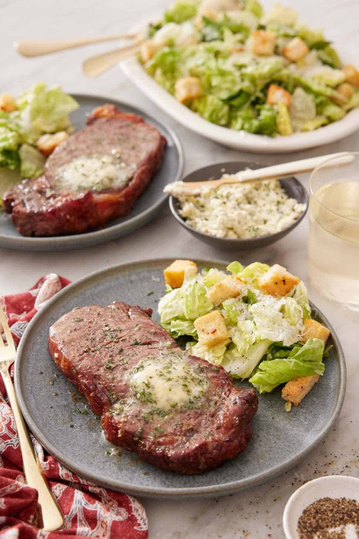 A plate with an air fryer steak with a side of salad. An additional plated serving in the background along with a platter of salad.