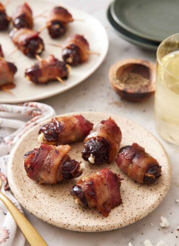 A small plate with five bacon wrapped dates with a platter in the background along with pepper and a drink.
