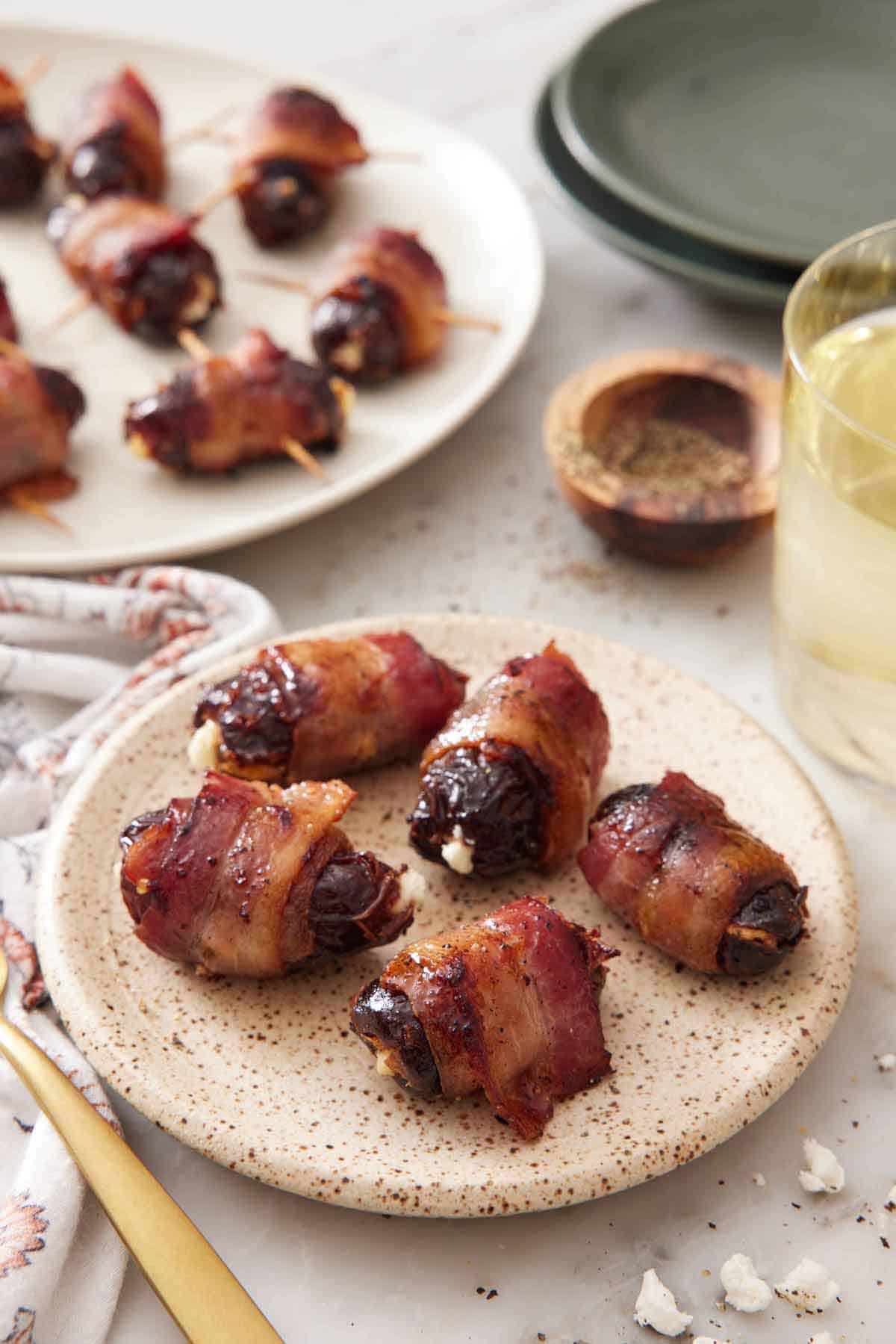 A small plate with five bacon wrapped dates with a platter in the background along with pepper and a drink.