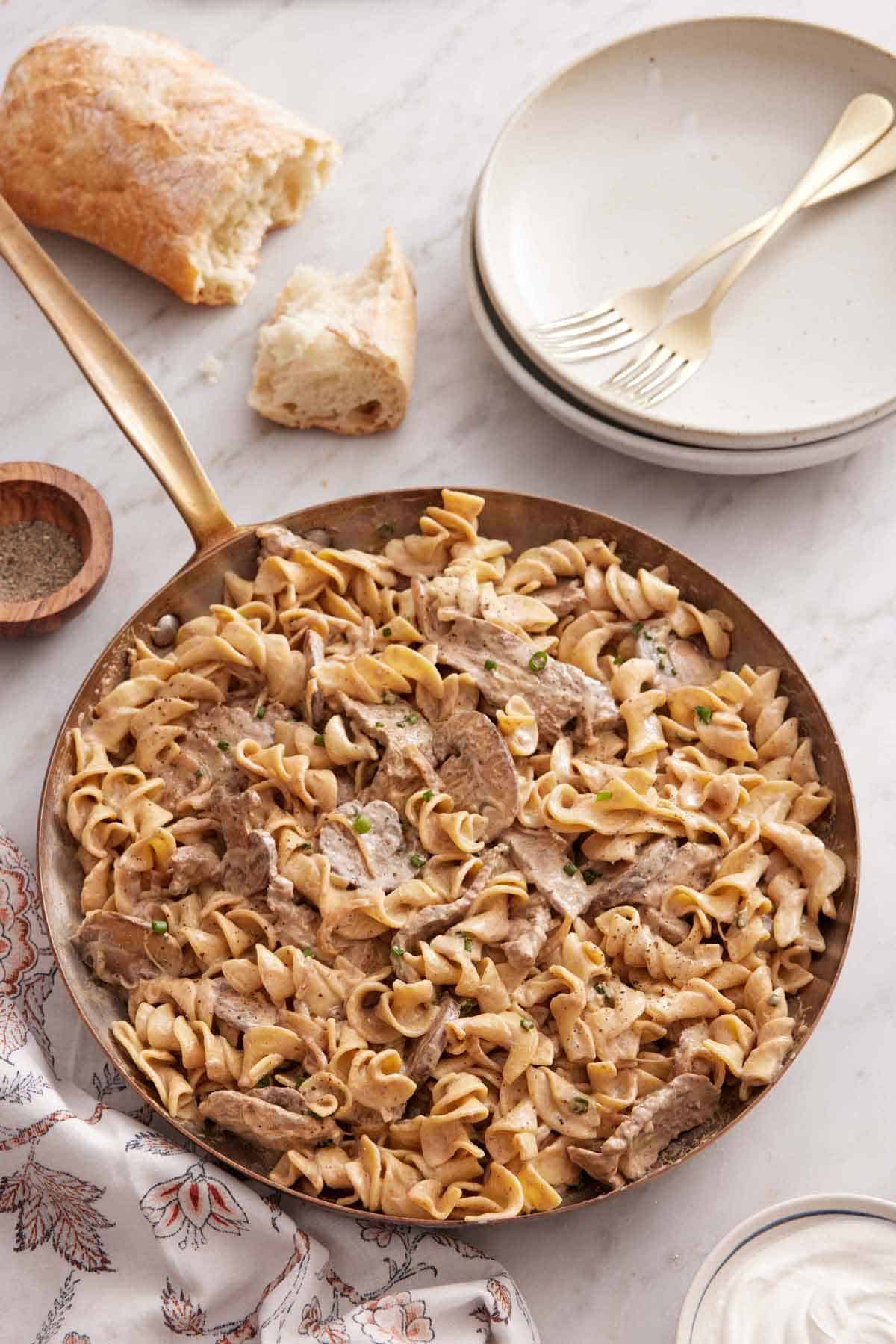 An overhead view of a skillet of beef stroganoff with some torn bread and a stack of plates with forks off to the side.