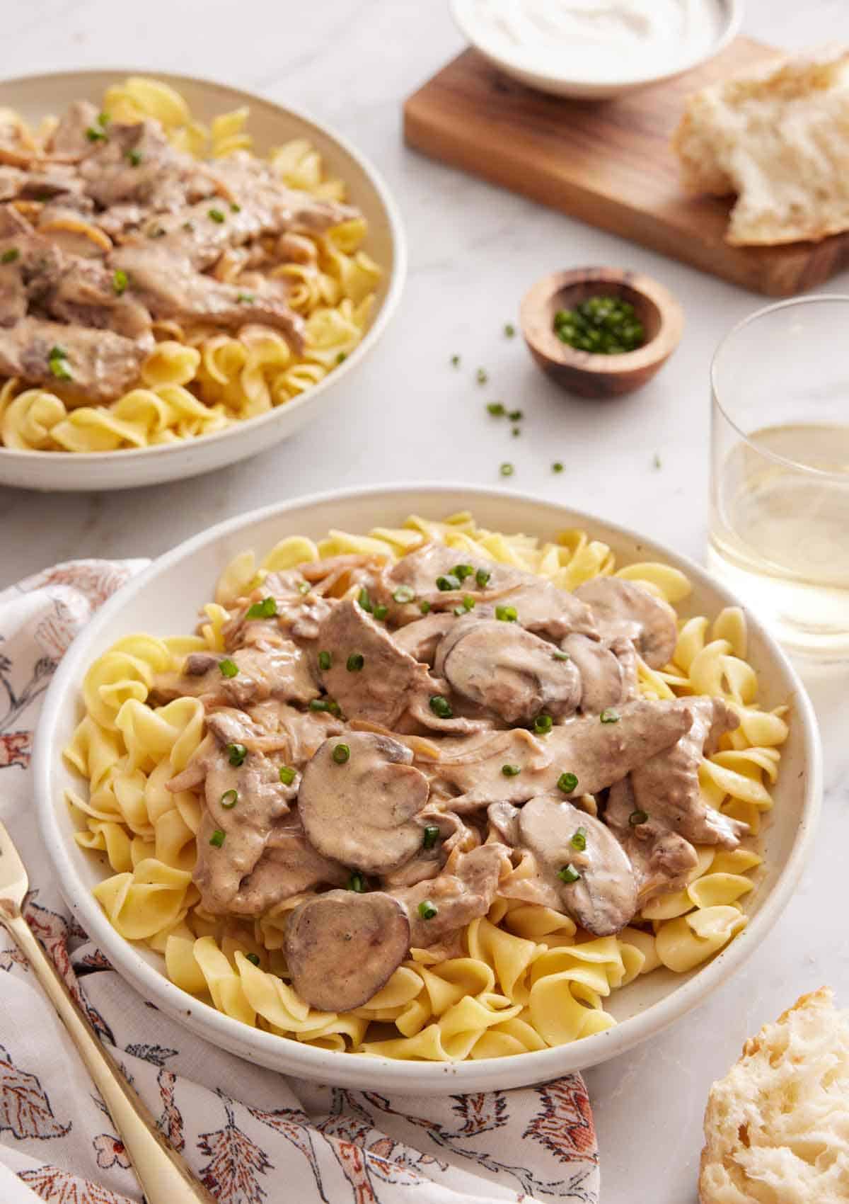 A plate of beef stroganoff topped with chives with a second plate, a drink, and torn bread in the background.