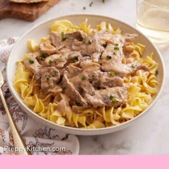 Pinterest graphic of a bowl of beef stroganoff topped with chives with a small bowl of chives, drink, and bread off to the side.
