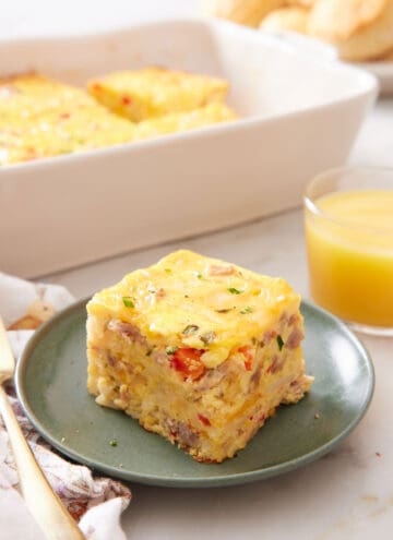 A plate with a serving of breakfast casserole with a baking dish and orange juice in the background.