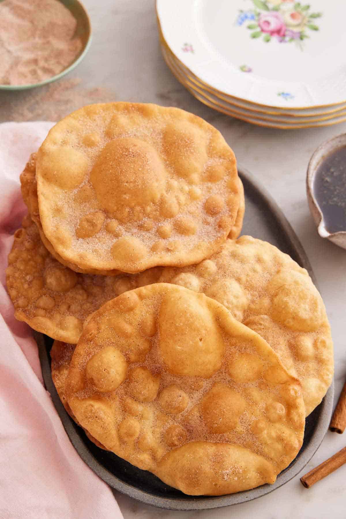 A platter with multiple pieces of bunuelos with a stack of plates in the background and cinnamon sugar.
