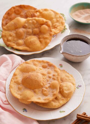A plate with two pieces of bunuelos with more in the background along with the piloncillo syrup.