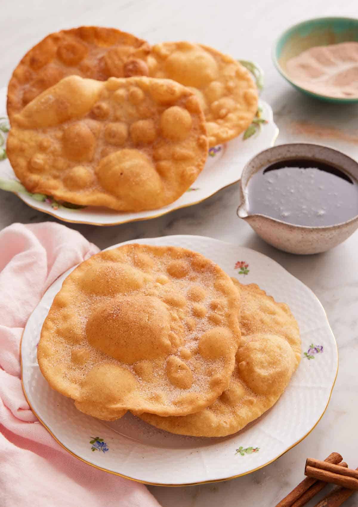 A plate with two pieces of bunuelos with more in the background along with the piloncillo syrup.