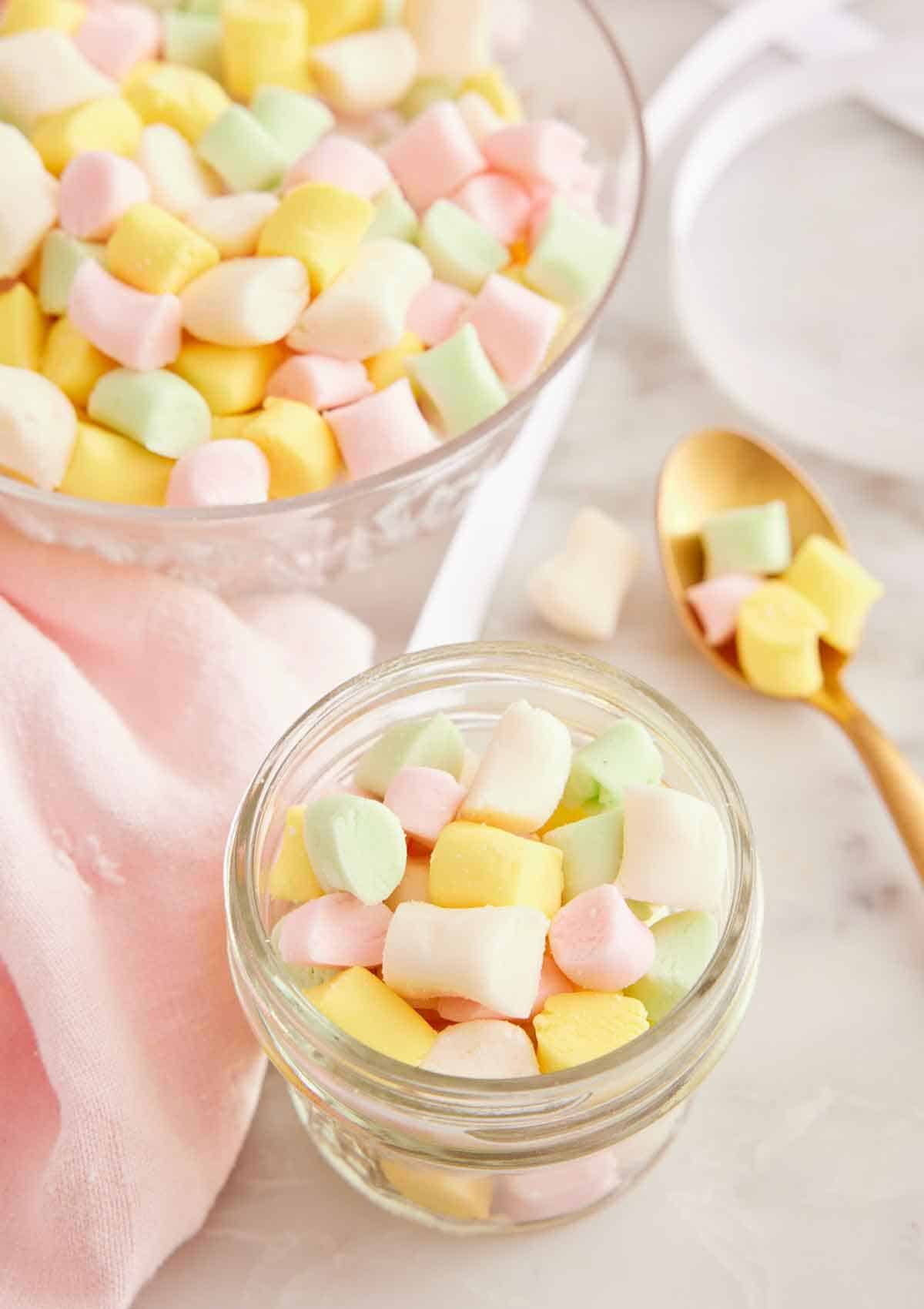 A mason jar with butter mints with a spoonful beside it and a bowl full in the background.