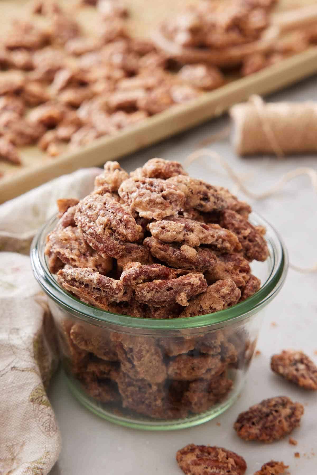 A jar of candied pecans with some on a sheet pan in the background.