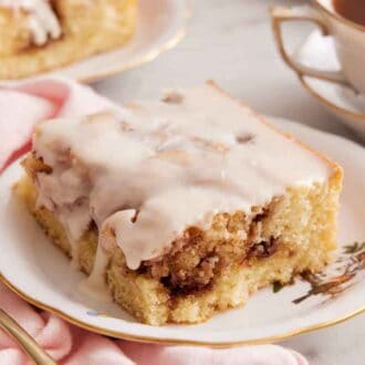 A plate with a slice of cinnamon roll cake with a cup of coffee in the back.