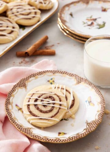 A plate with two cinnamon roll cookies along with a glass of milk, stack of plates, and more cookies in the background.