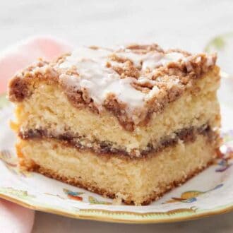 A piece of coffee cake on a plate topped with glaze. Cup of coffee in the background.