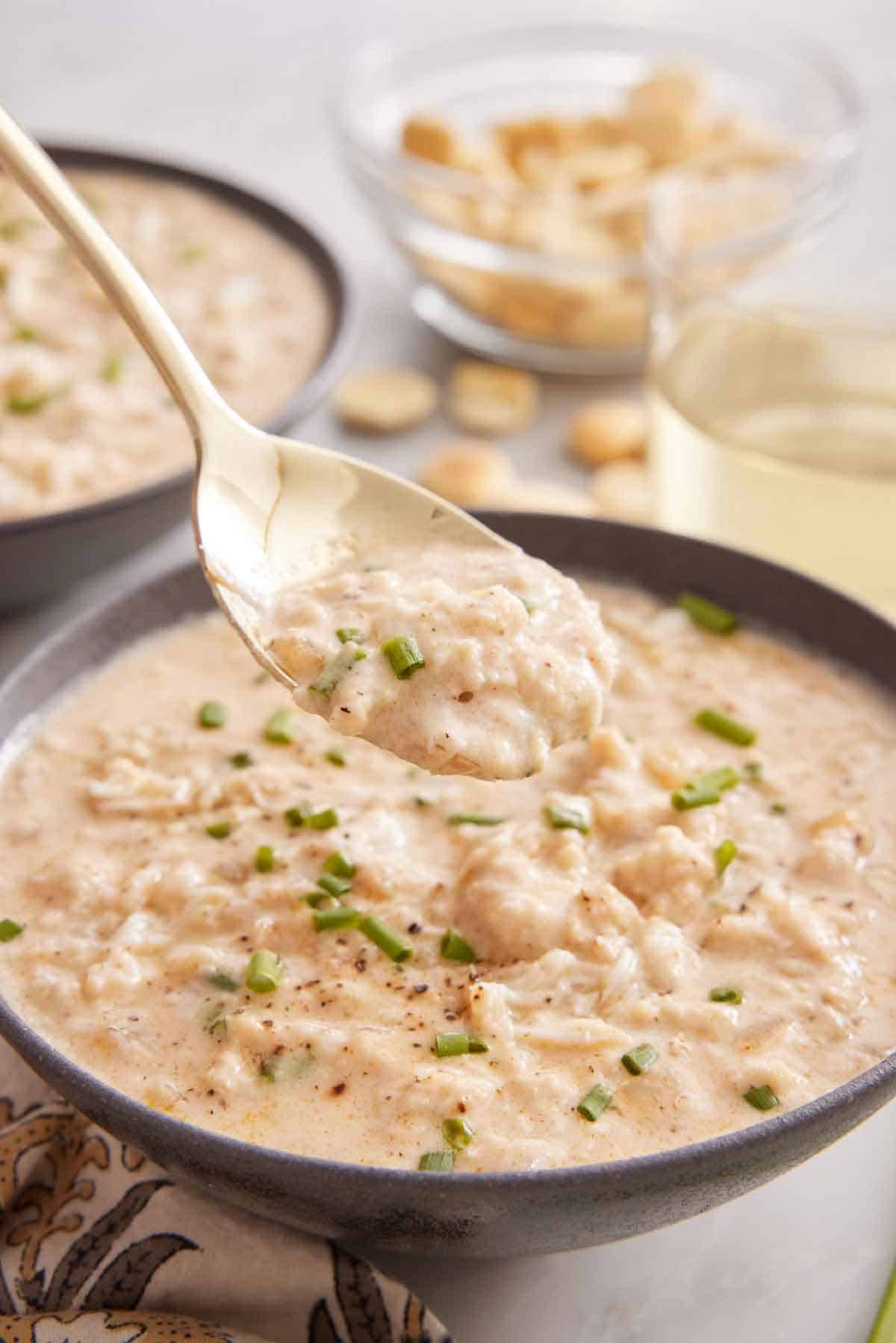 A spoonful of crab soup lifted from the soup bowl. A drink in the back along with more crackers.