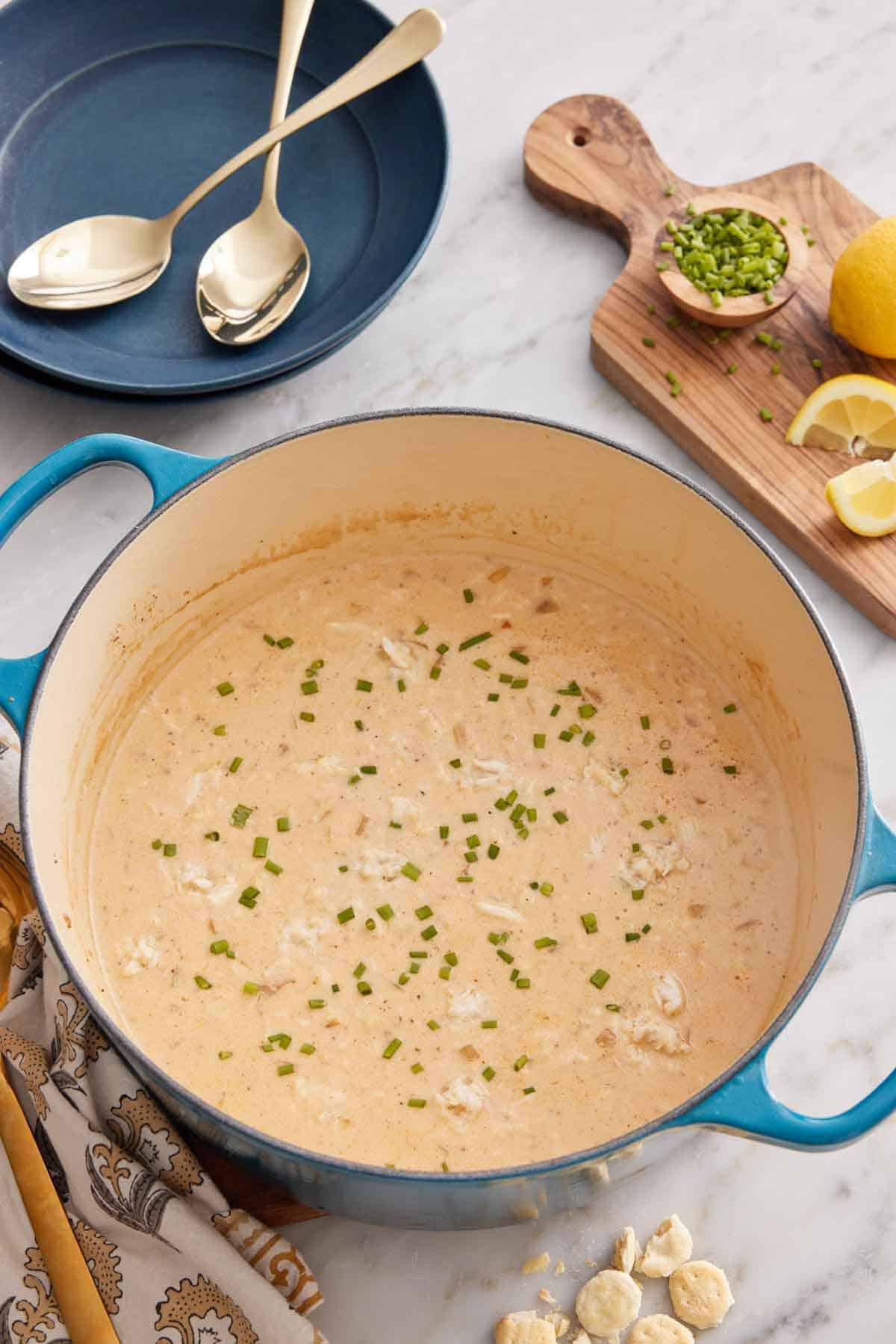 An overhead view of a pot of crab soup. Garnishes on a serving board and a bowl with two spoons beside it.