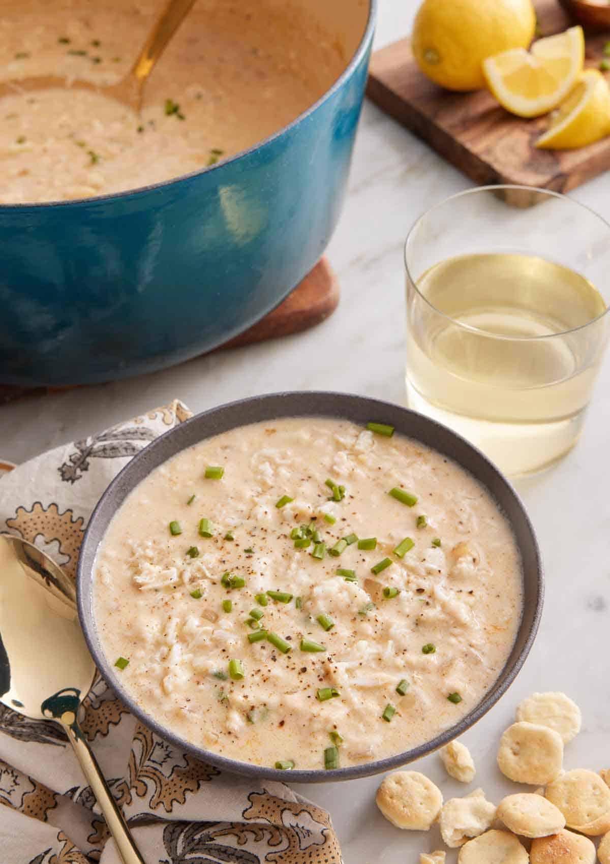 A bowl of crab soup with a drink and pot with the rest of the soup in the background.