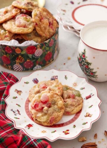A plate with three fruitcake cookies with a tin in the background and a cup of milk.