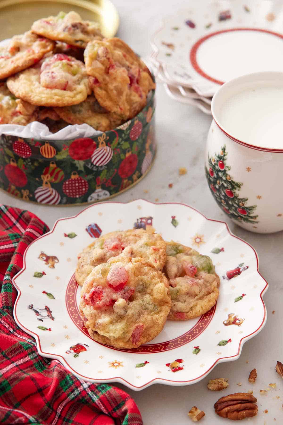 A plate with three fruitcake cookies with a tin in the background and a cup of milk.