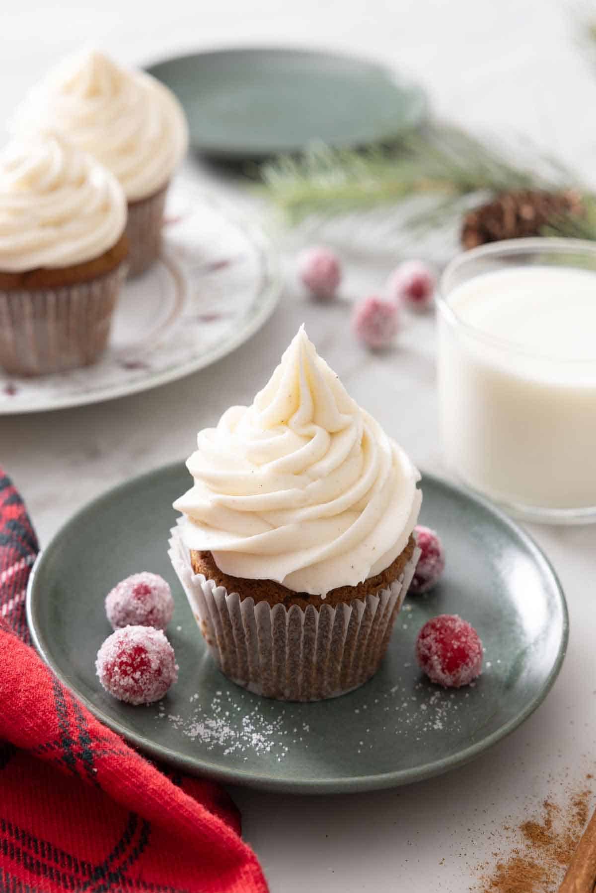 A plate with a gingerbread cupcake topped with cream cheese frosting. Two more cupcakes in the background and a glass of milk.