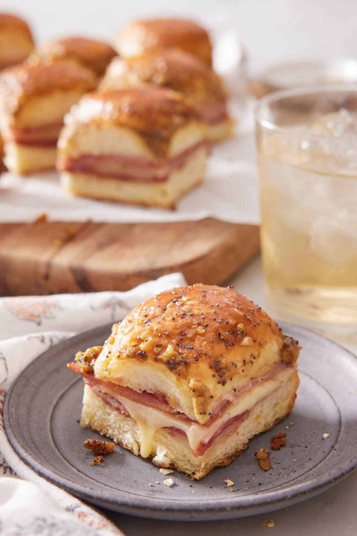 A plate with a ham and cheese slider with an iced drink and more sliders in the back.