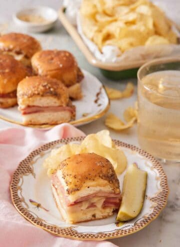 A plate with a ham and cheese slider along with chips and a pickle. More sliders, chips, and a drink in the background.