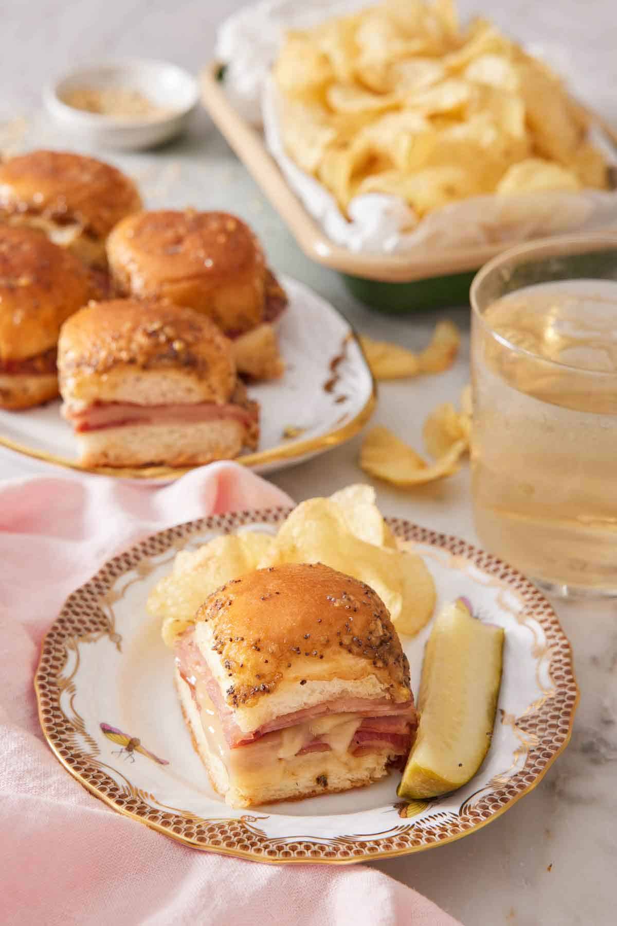 A plate with a ham and cheese slider along with chips and a pickle. More sliders, chips, and a drink in the background.