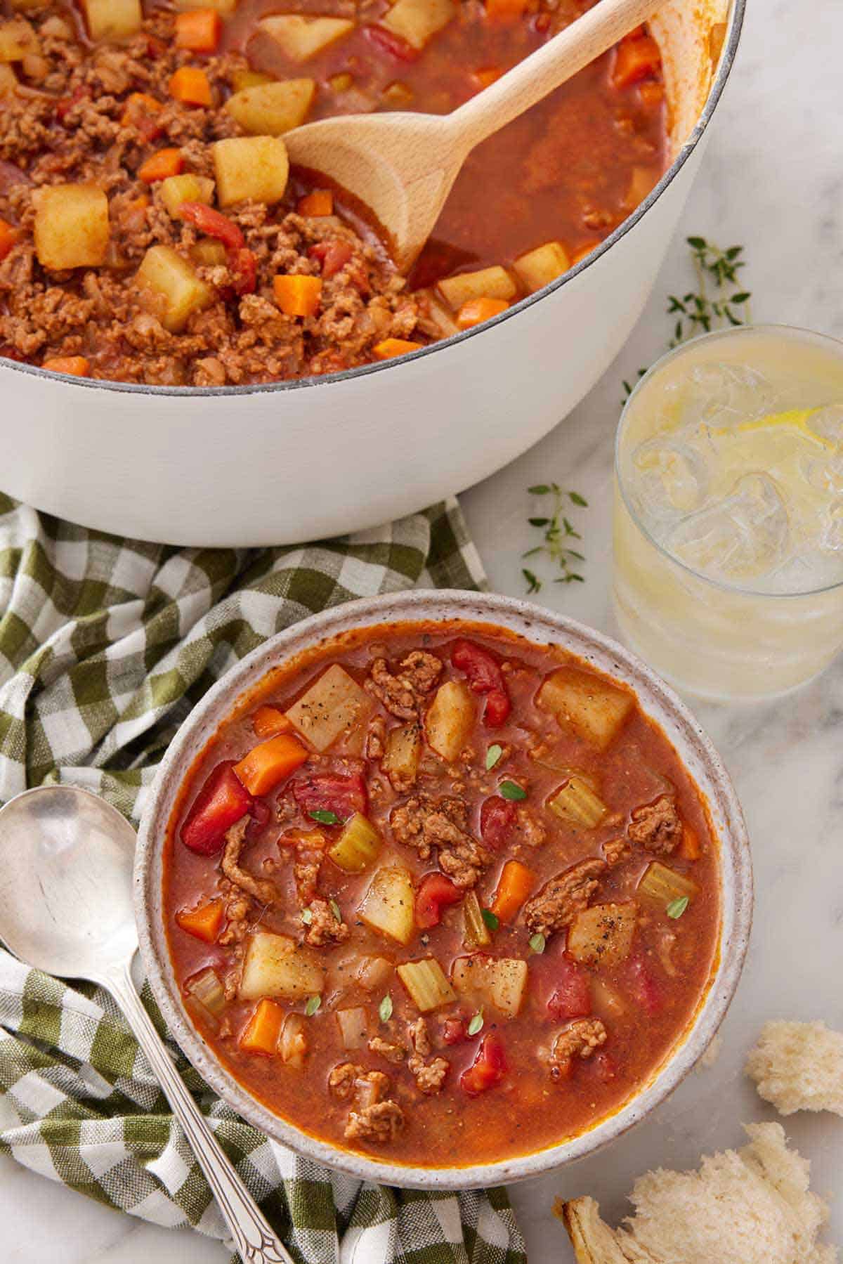 Overhead view of a bowl of hamburger soup beside a drink and the rest of the soup in a pot.