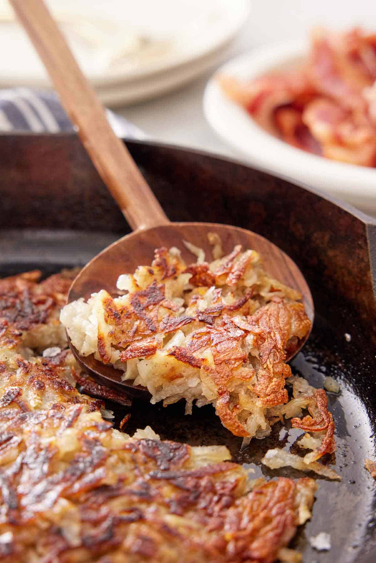 A wooden spoon lifting hash browns off a cast iron skillet.