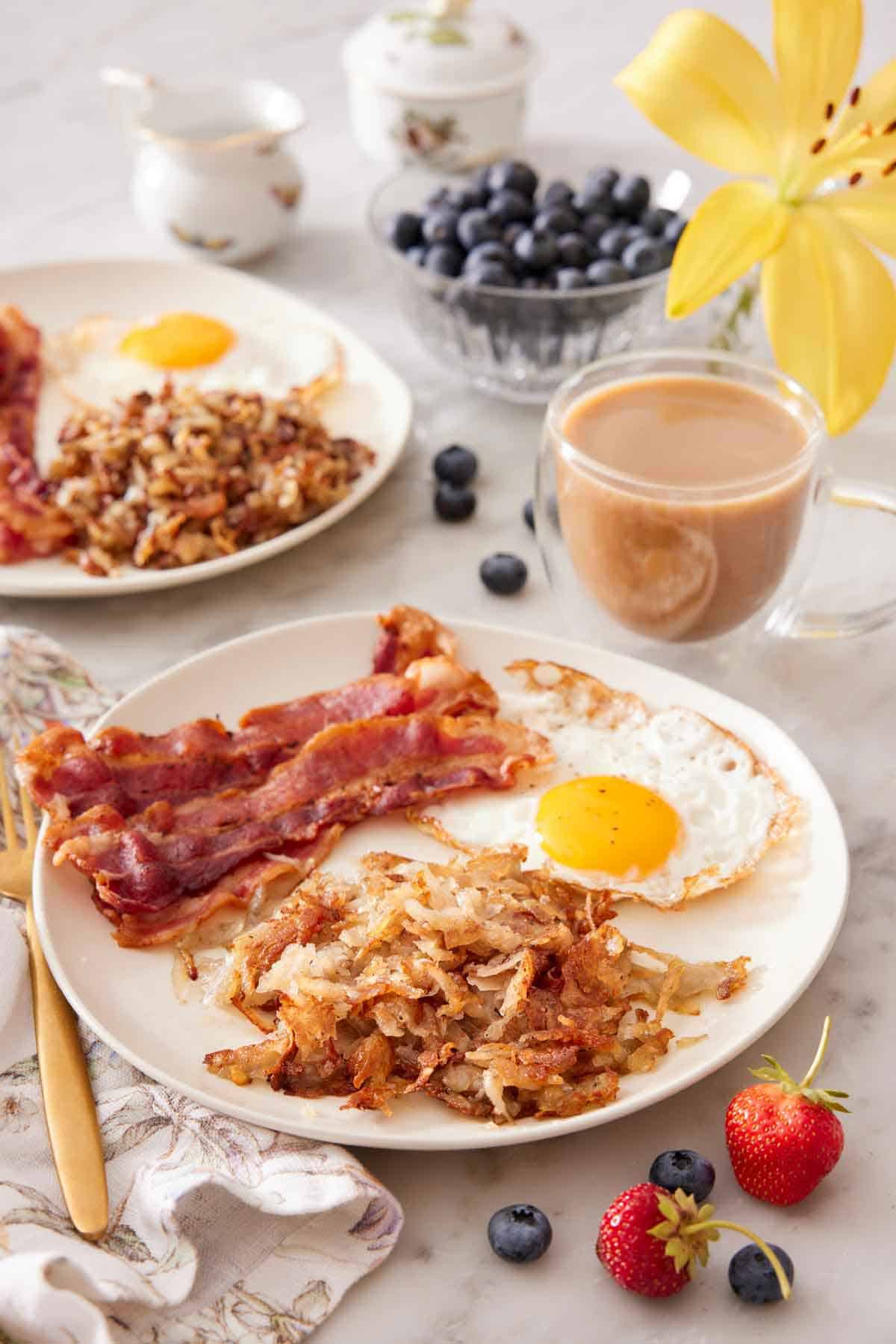 A plate with hash browns, bacon strips, and a fried egg with a cup of coffee, bowl of blueberries, and additional plate in the background.