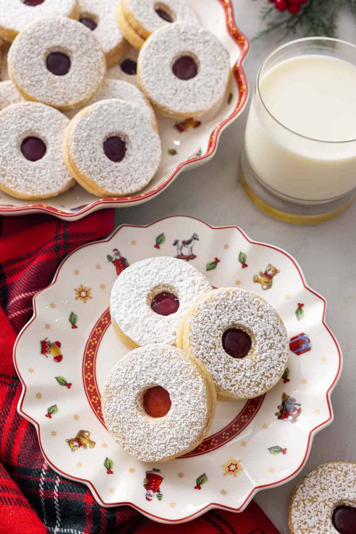 A festive plate with three linzer cookies by a mug of milk and a platter of cookies.