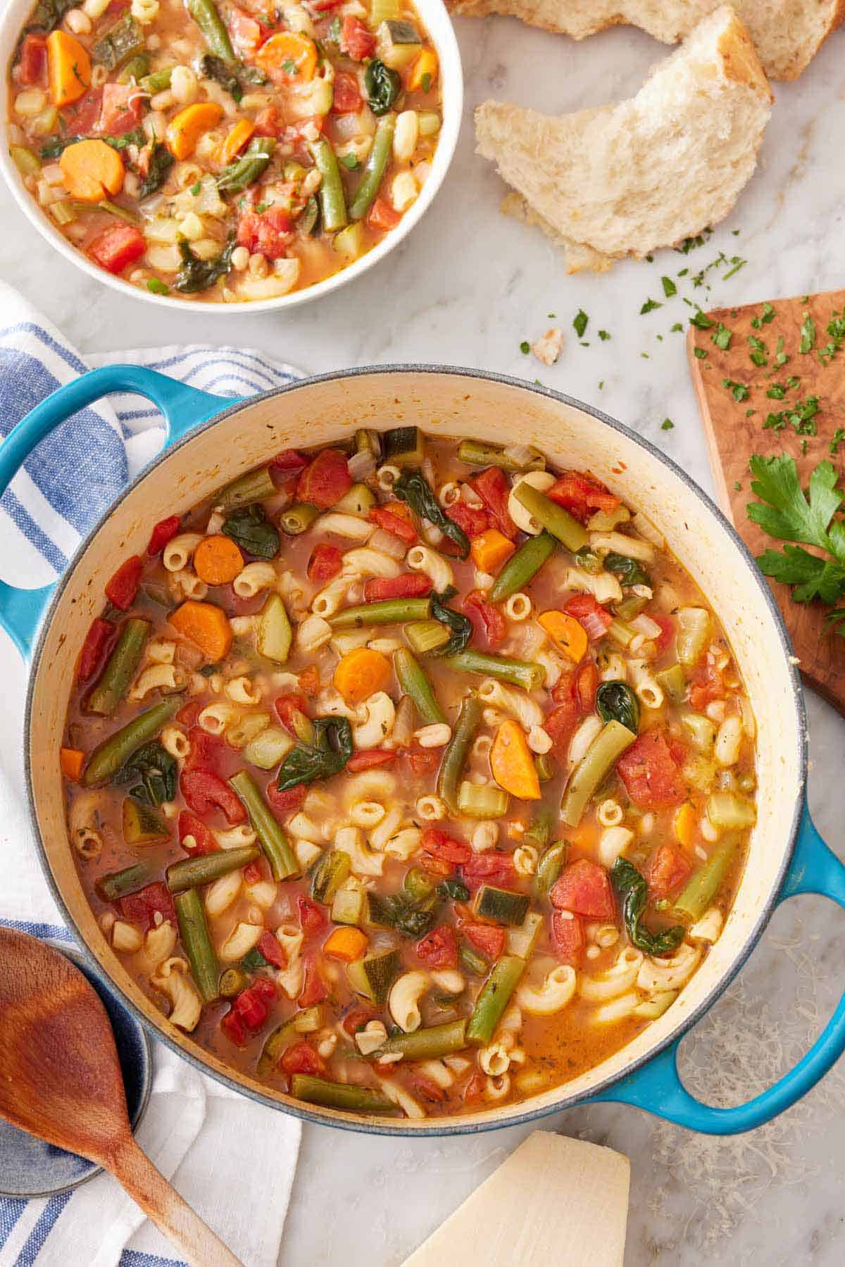 Overhead view of a pot of minestrone soup with a bowl of soup and some torn bread off to the side.