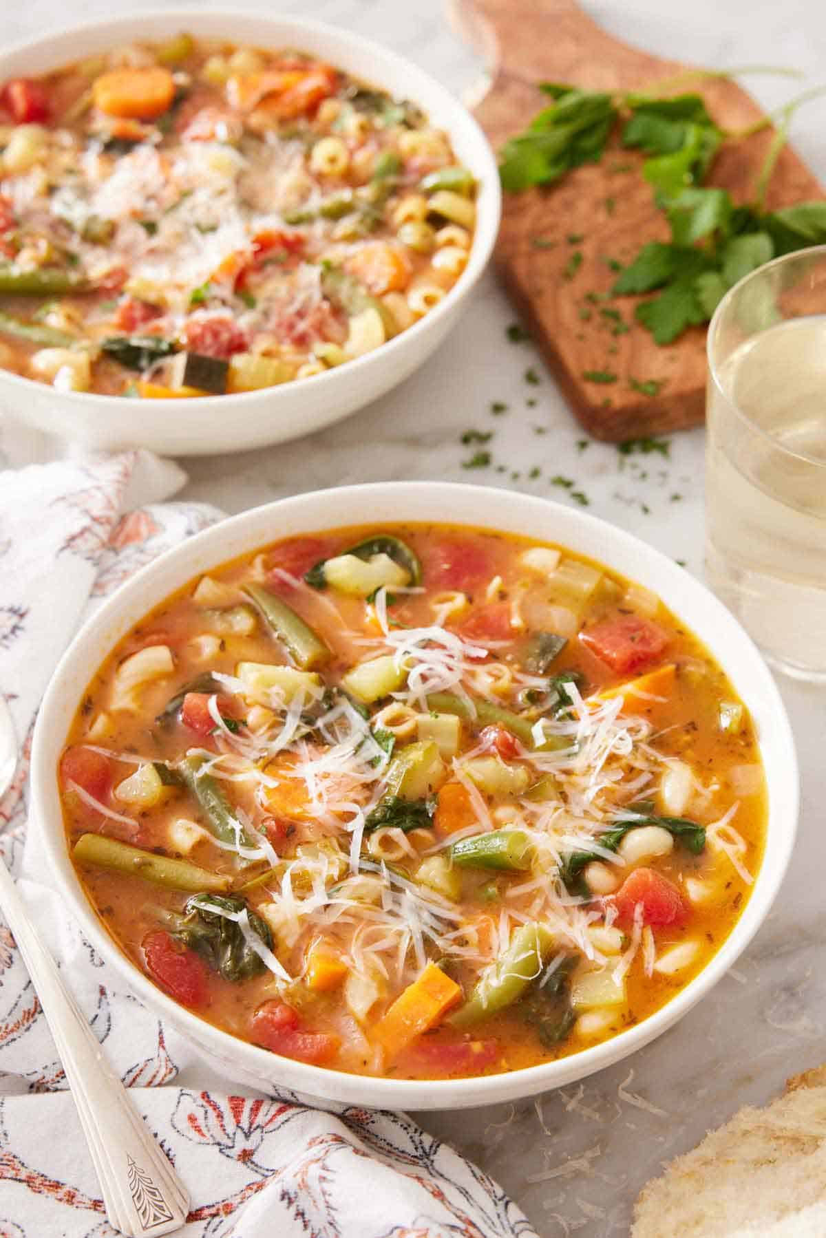 Two bowls of minestrone soup with one in front, topped with parmesan cheese. Parsley and a glass of wine in the background.