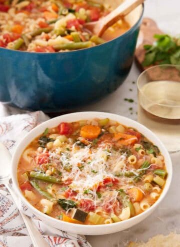 A bowl of minestrone soup with a glass of wine and pot of soup in the background.