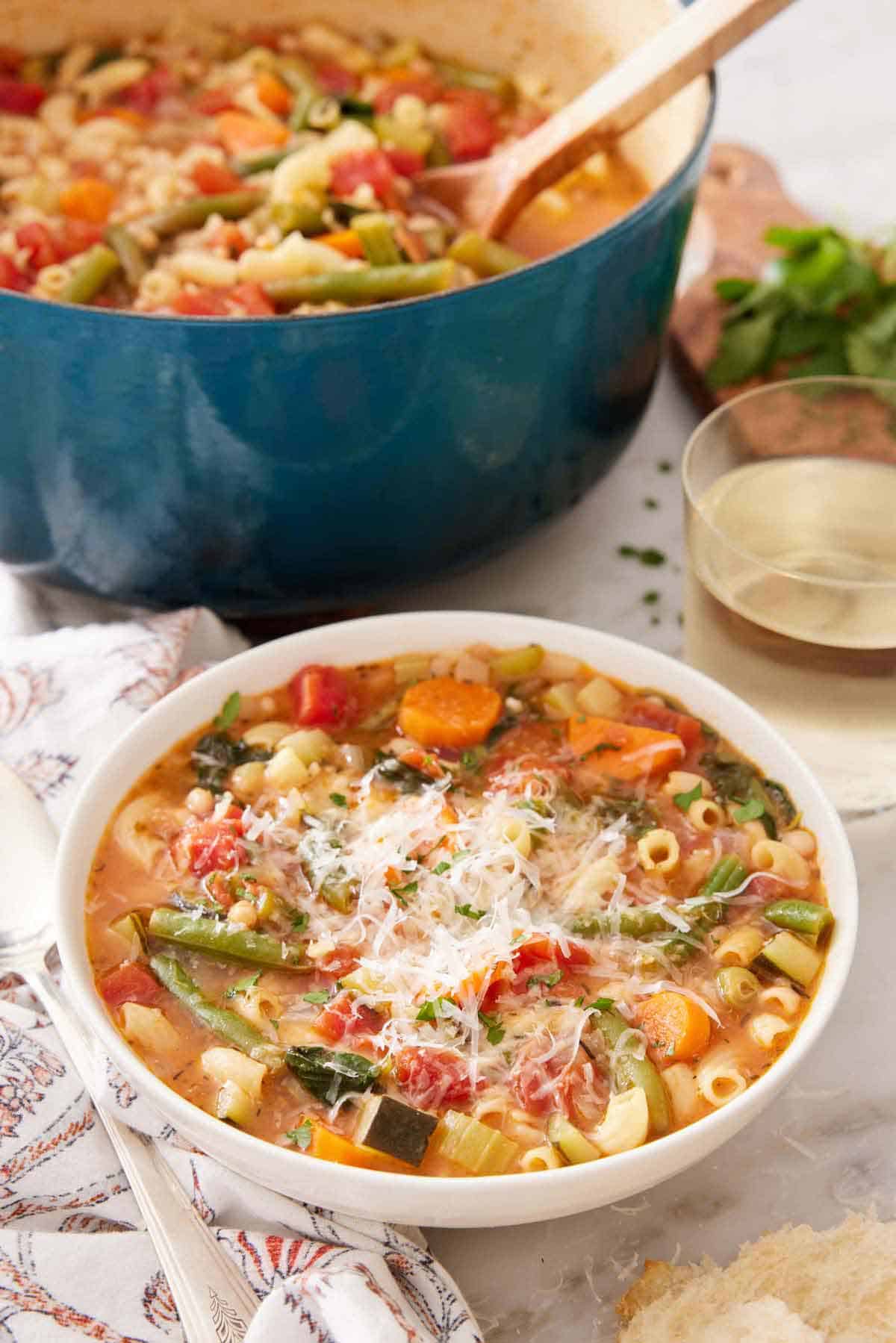 A bowl of minestrone soup with a glass of wine and pot of soup in the background.